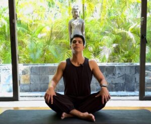 Mauricio from Livana Life demonstrating a seated forward fold yoga stretch to release neck and shoulder pain, sitting on a dark mat in a calm studio environment